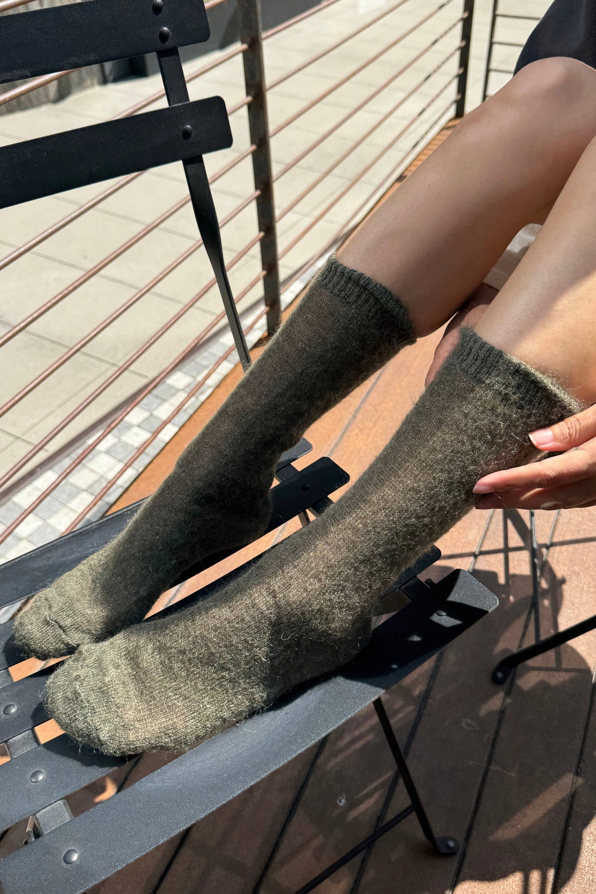 A person wearing fuzzy ribbed knee-high socks in a muted olive-brown tone, seated on a black metal chair in an outdoor setting.