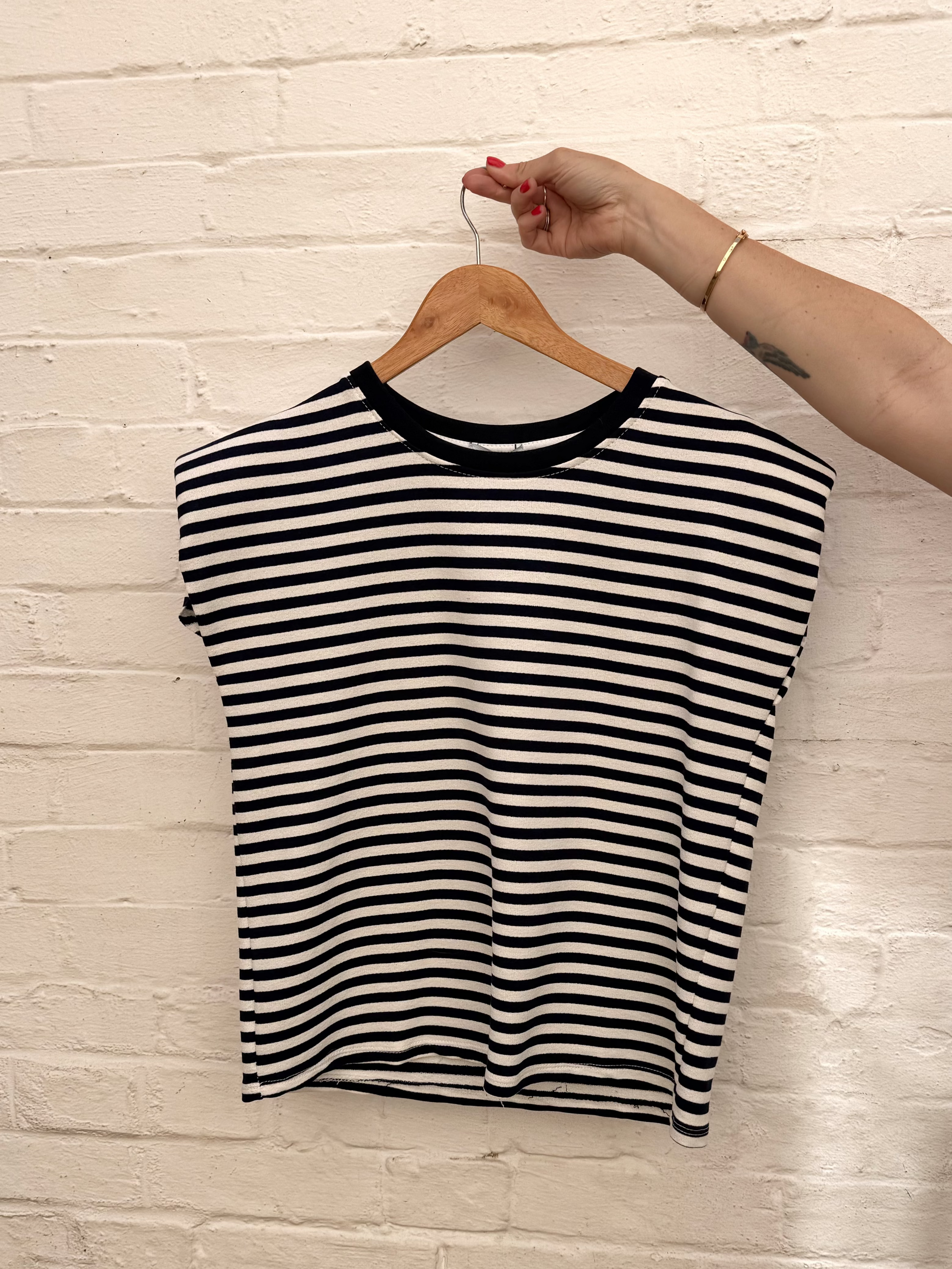 A black and white horizontally striped short-sleeve crop top with a black crew neck, hanging on a wooden hanger held by a person's hand against a white brick wall.