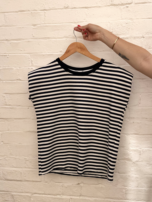 A black and white horizontally striped short-sleeve crop top with a black crew neck, hanging on a wooden hanger held by a person's hand against a white brick wall.