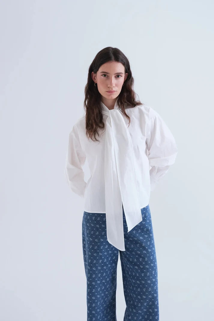A woman modeling a white long-sleeve blouse with a sheer, oversized front bow and blue patterned trousers, set against a plain light gray studio background.