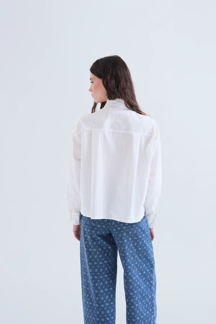 A woman shown from the back wearing a white long-sleeve button-up shirt and blue patterned pants against a plain white studio background.