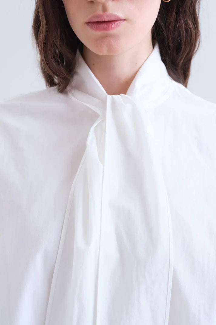 A close-up of a model wearing a white high-neck blouse with a large front bow and tie detail, set against a clean white background.