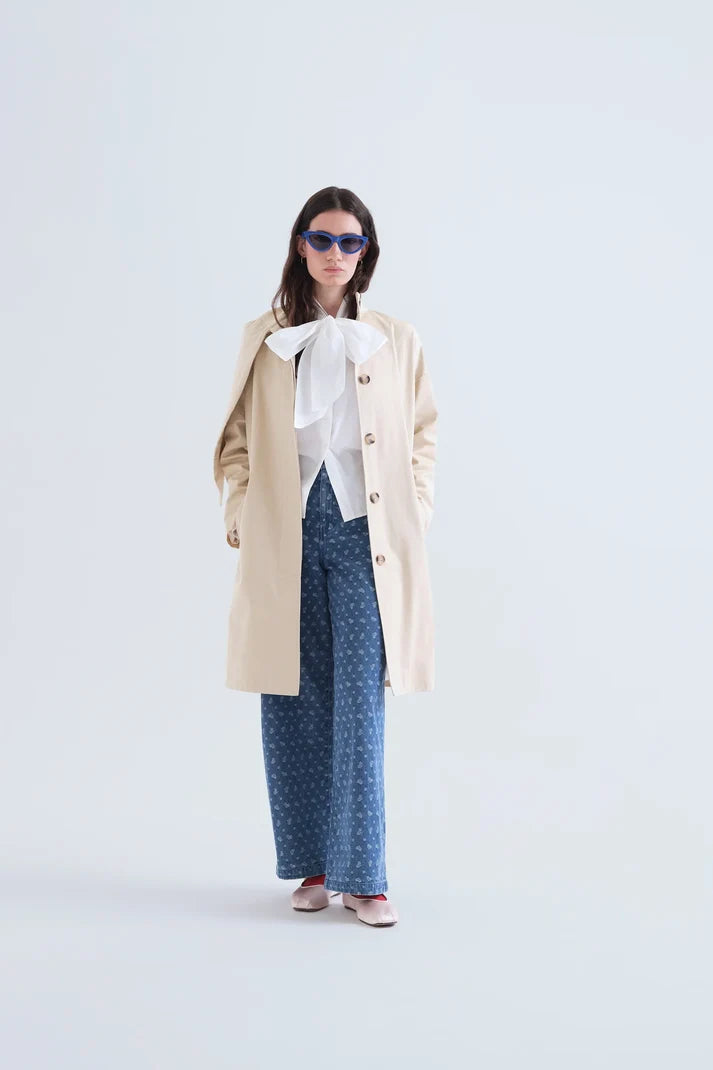 A model stands against a light gray studio background wearing a long cream coat over a white blouse with a large bow, blue wide-leg patterned pants, and blue sunglasses.