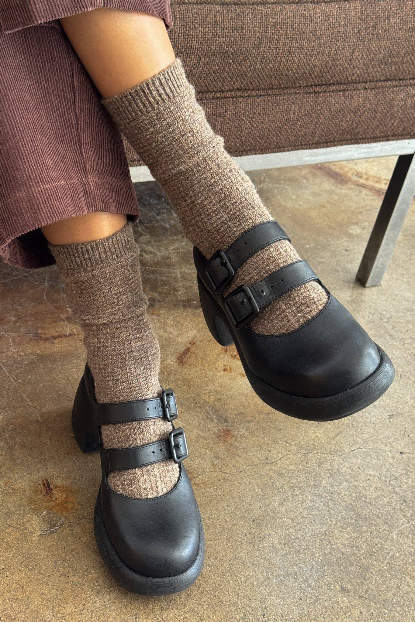 Black clogs with brown socks worn on a concrete floor.