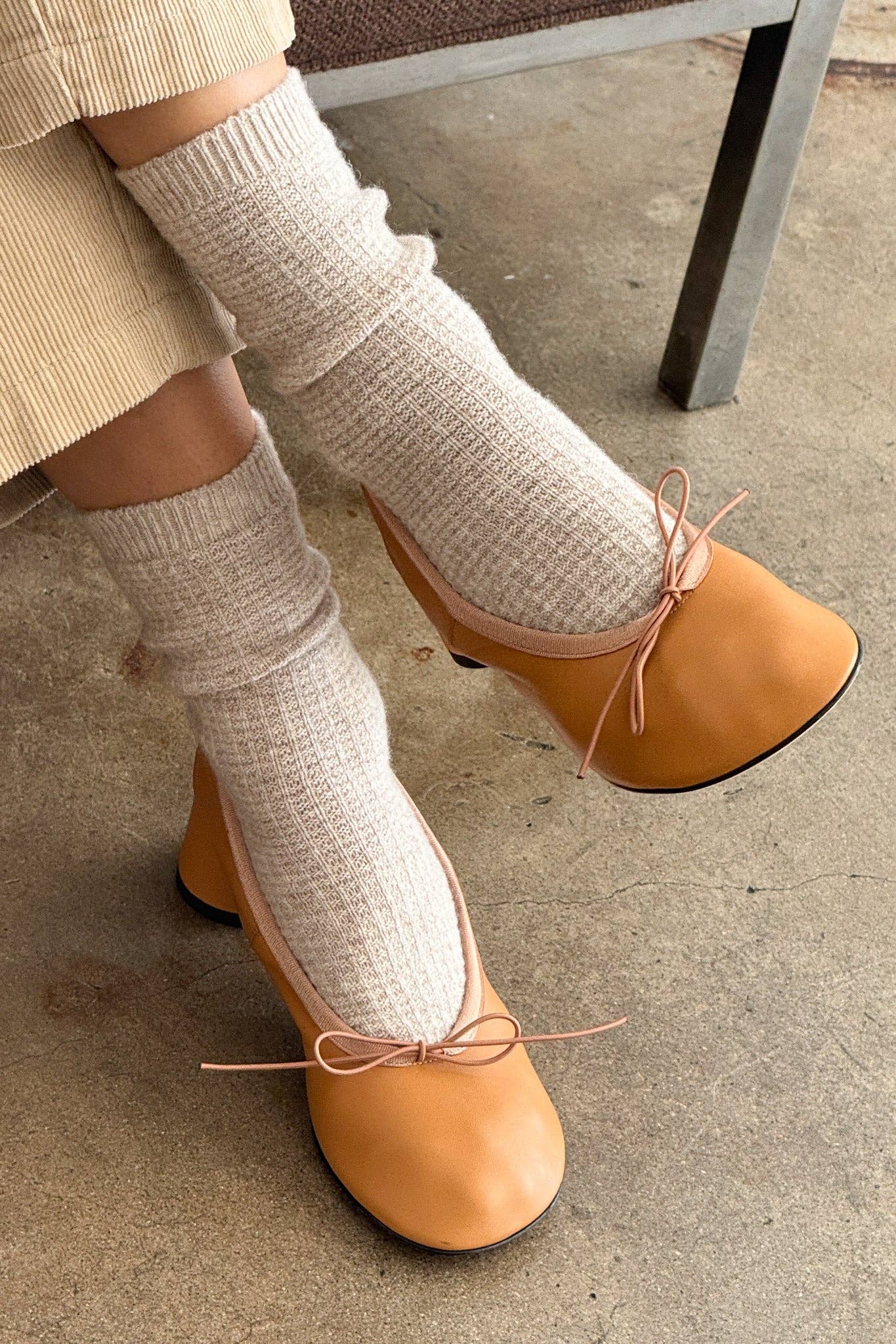 Person wearing tan shoes with bow details and white leg warmers on a concrete floor.